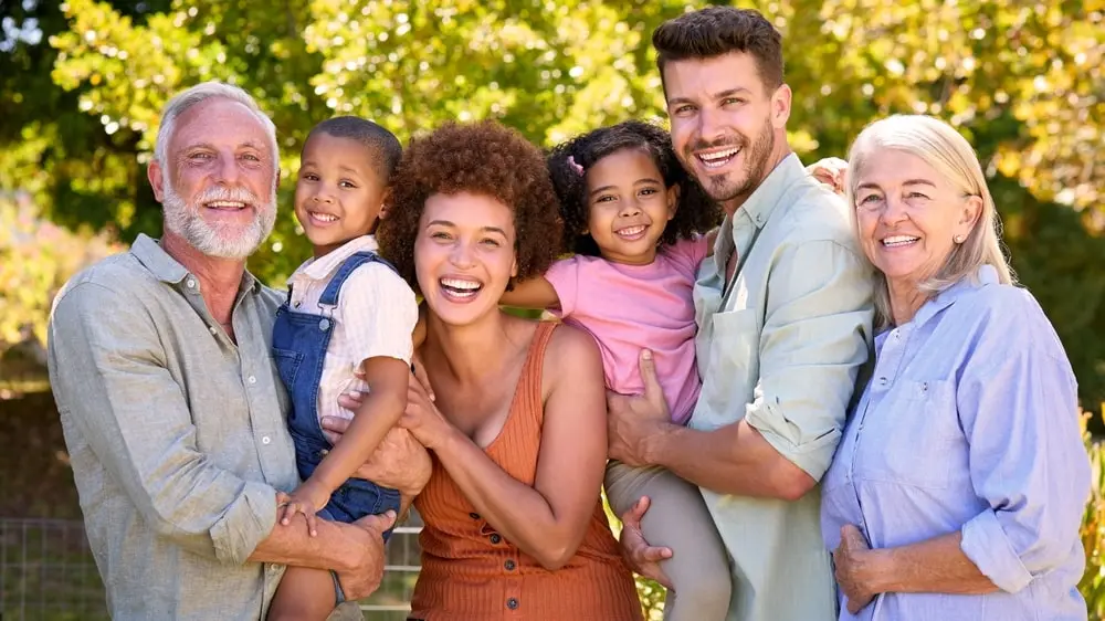 A joyful family of six poses outdoors, including two senior adults, two young adults, and two children. They are smiling in a sunny park setting with trees in the background, all embracing each other warmly.