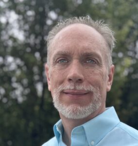A middle-aged man with light skin, short gray hair, and a trimmed beard stands outdoors, wearing a light blue collared shirt. Trees with green leaves are blurred in the background.