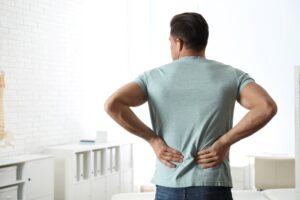 A man in a light blue t-shirt stands indoors with his back to the camera, holding his lower back with both hands, suggesting he may be experiencing back pain.