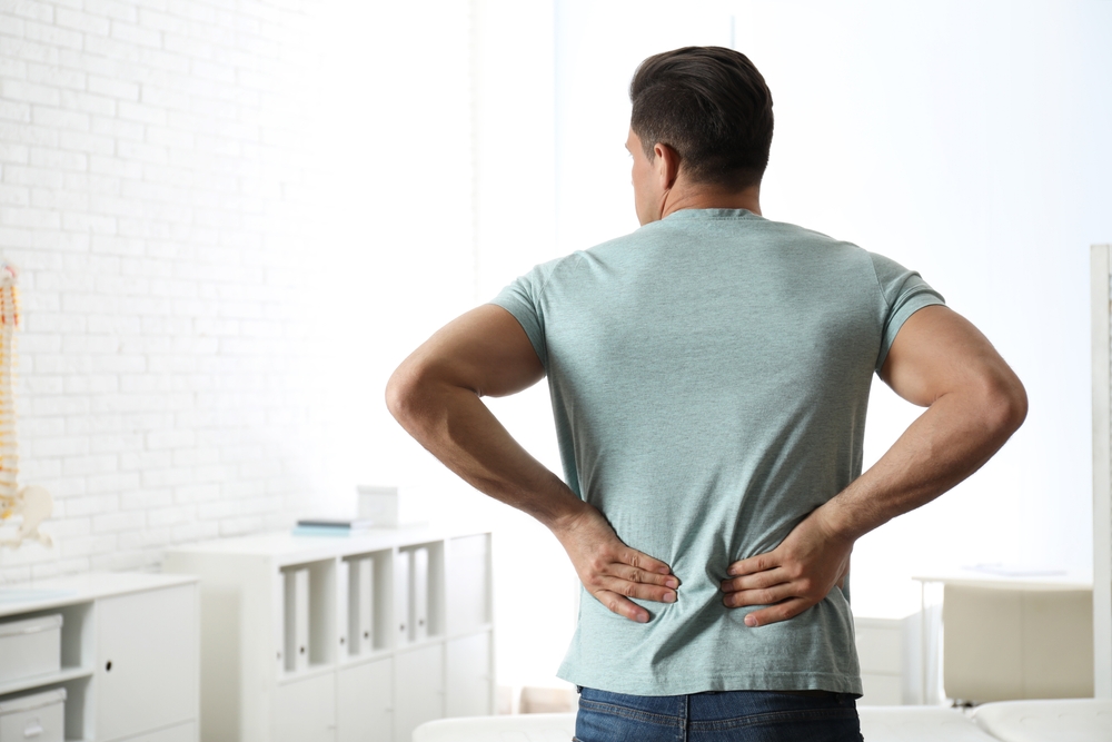 A man in a light blue t-shirt stands indoors with his back to the camera, holding his lower back with both hands, suggesting he may be experiencing back pain.
