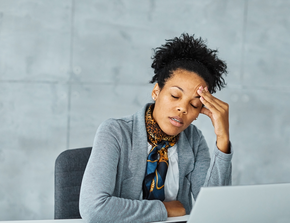 A woman sits at a desk with her eyes closed, resting her head on one hand and looking tired or stressed. She is wearing a gray blazer and patterned scarf, with a laptop in front of her.