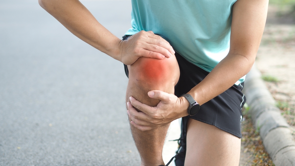 A person wearing a light blue shirt and black shorts is kneeling on one knee outdoors, holding their knee with both hands. The knee appears red, indicating pain or injury.