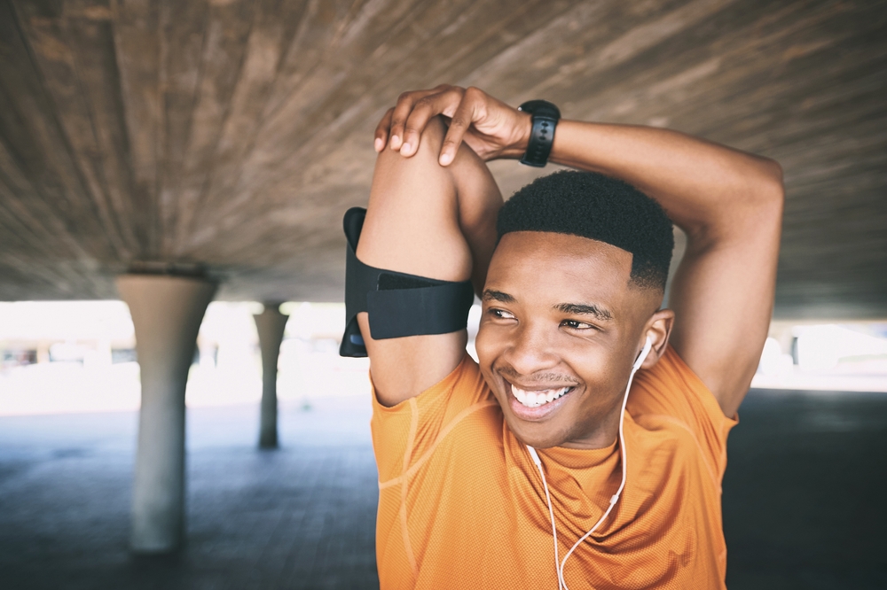 A young man in an orange shirt stretches his arm overhead while smiling. He wears earphones, a smartwatch, and an armband, standing under a structure with concrete pillars.