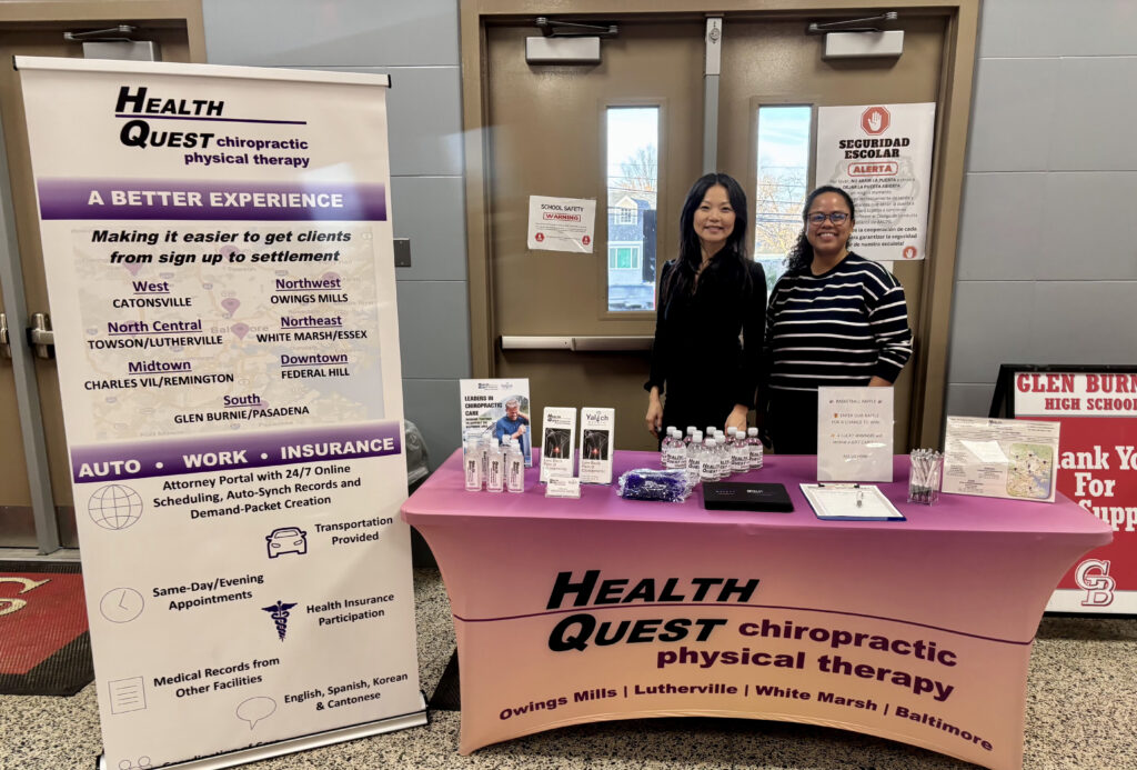 Two women stand behind a pink Health Quest Chiropractic Physical Therapy table with brochures, water bottles, and pens. A large informational banner is displayed on the left, and gray double doors are in the background.
