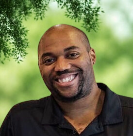 A smiling man with a shaved head and short beard wearing a black collared shirt stands outdoors with green foliage and blurred trees in the background.