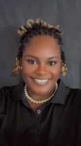 A woman with styled hair in twists, wearing a black collared shirt and a pearl necklace, smiles at the camera against a plain dark background.