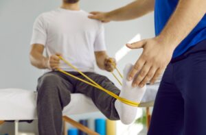 A person sits on an exam table, stretching their foot with a yellow resistance band, while a therapist in blue guides and supports them during a physical therapy session.