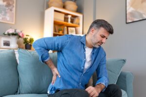 A man sitting on a couch holds his lower back and appears to be in pain, suggesting discomfort or back pain. The room has soft blue decor, shelves, and a vase of flowers in the background.